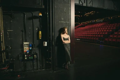 Model standing backstage leaning against a dark wall beside an empty theatre auditorium with red seats