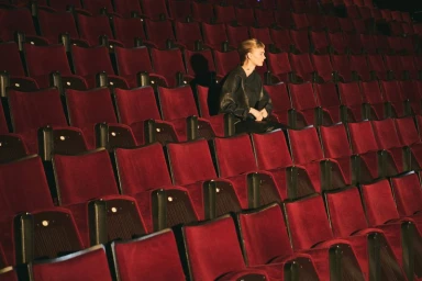 Model Caitlin Soetendal seated alone wearing a dark jacket surrounded by rows of empty red velvet auditorium chairs.