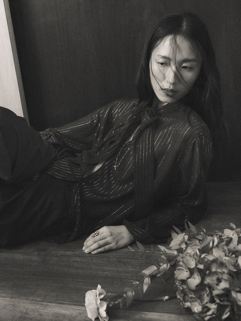 Monochrome portrait of an Asian woman in a sheer blouse, lying on a table next to flowers, with dark backdrop and soft, diffused lighting.