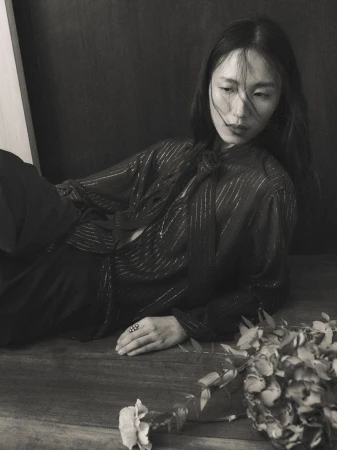 Monochrome portrait of an Asian woman in a sheer blouse, lying on a table next to flowers, with dark backdrop and soft, diffused lighting.