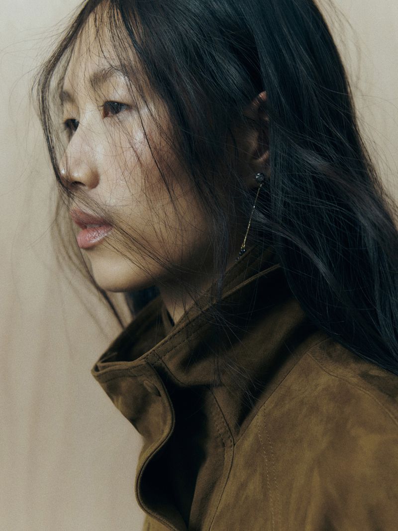 Portrait of an Asian woman with long dark hair, wearing a brown jacket and single earring against a neutral background in soft lighting.