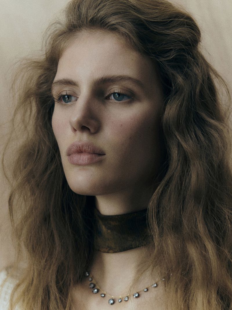 Close-up portrait of a fair-skinned woman with wavy brown hair wearing a brown velvet choker and delicate beaded necklace looking downward.