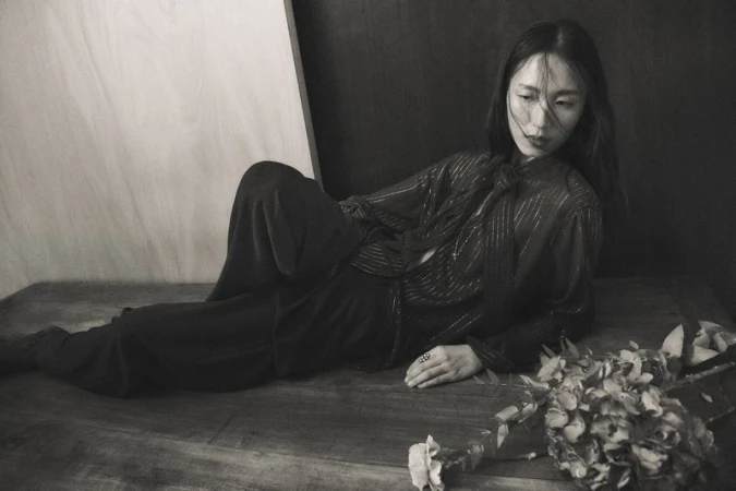Monochromatic portrait of an adult female lying on wooden surface next to flowers, wearing a patterned blouse and dark pants.