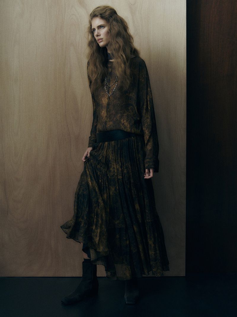 Model with long wavy hair posing in a dark bohemian printed blouse and pleated maxi skirt against a light wood paneled wall.
