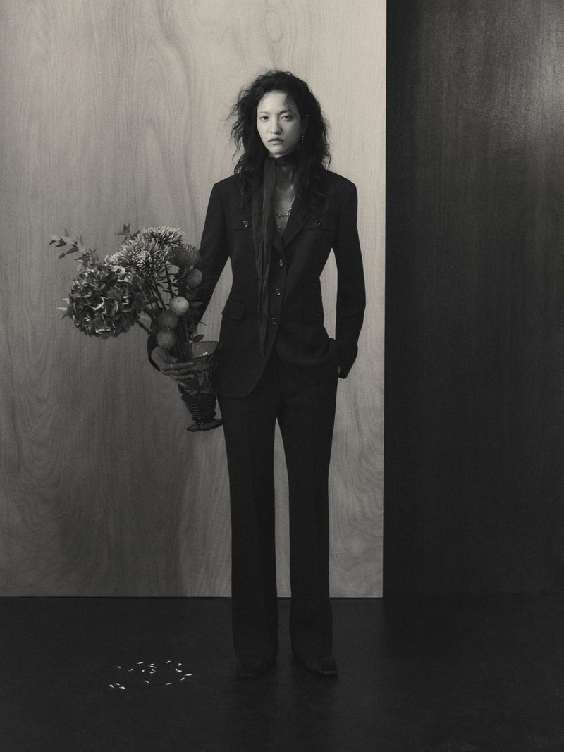 Monochrome portrait of a stylish Asian woman in a black suit holding a vase of flowers against a neutral plywood and dark backdrop.