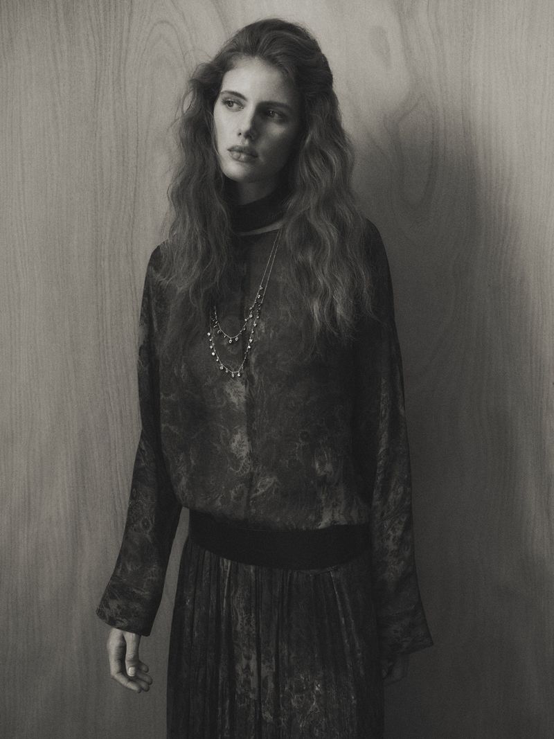 Monochrome portrait of a young woman with long, wavy hair wearing a patterned top and skirt against a neutral background.