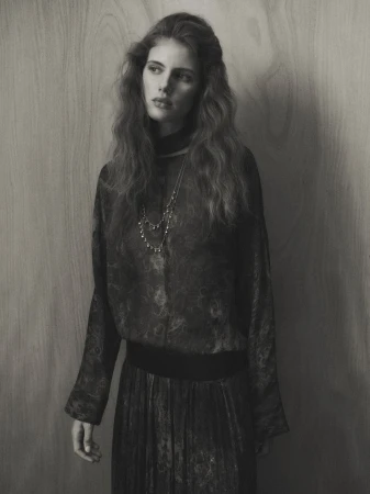 Monochrome portrait of a young woman with long, wavy hair wearing a patterned top and skirt against a neutral background.