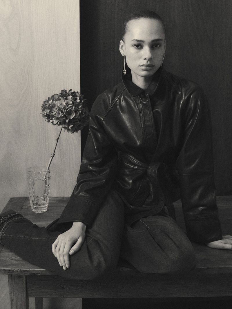 Monochromatic shot of a woman wearing a leather jacket sitting on a wooden surface with a flower in a glass vase beside her.