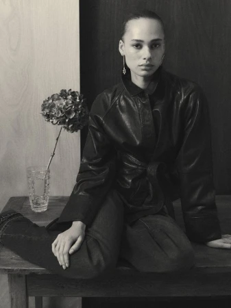 Monochromatic shot of a woman wearing a leather jacket sitting on a wooden surface with a flower in a glass vase beside her.