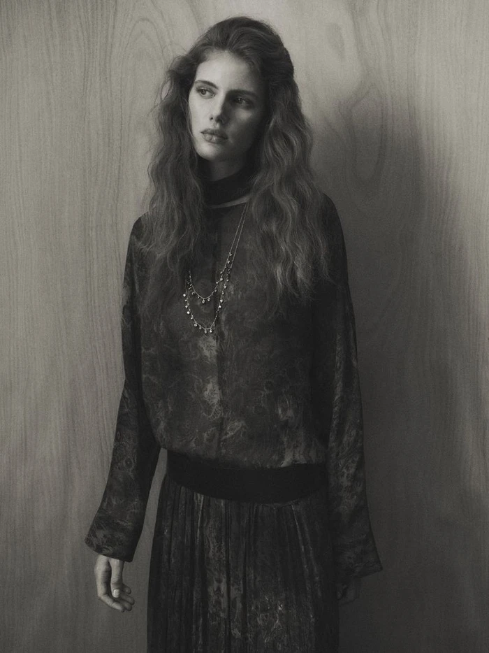 Model with long wavy hair wearing a patterned long-sleeve dress and layered necklace against a wood panel wall.