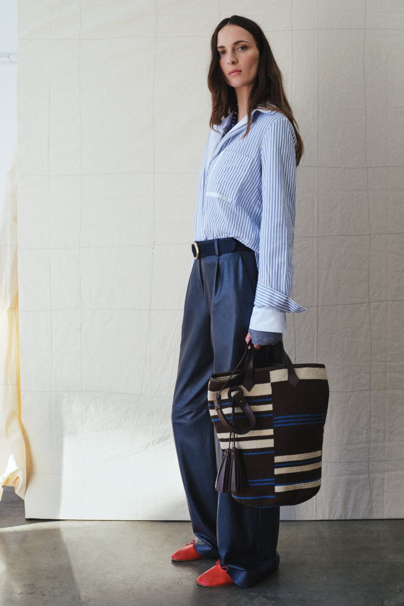 Stylish woman in a striped shirt, wide-leg pants, and red shoes poses with a striped tote bag against a neutral background.