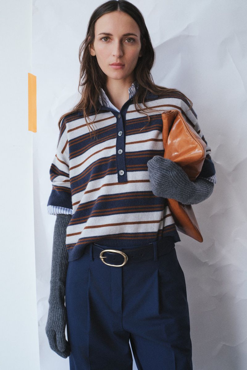 Fashionable woman posing in striped shirt, blue pants, and grey gloves with a tan clutch, set against a textured white backdrop.