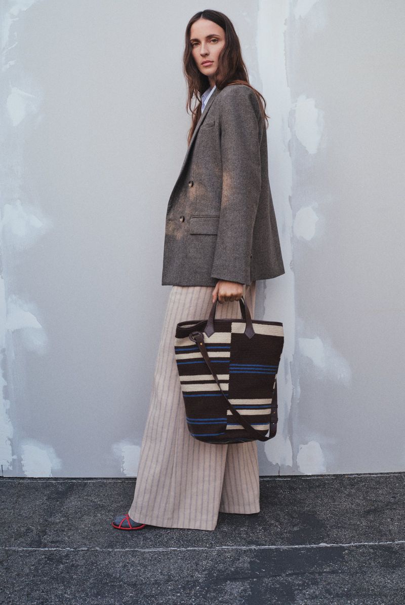 A woman stands in a grey herringbone blazer and striped trousers, holding a striped tote bag against a neutral plaster wall.