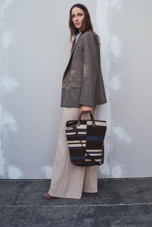 A woman stands in a grey herringbone blazer and striped trousers, holding a striped tote bag against a neutral plaster wall.