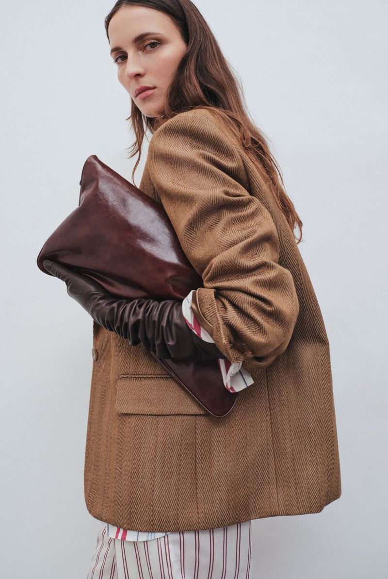 A woman in a brown herringbone blazer holds a shiny brown handbag with leather gloves against a light background in a fashion editorial.