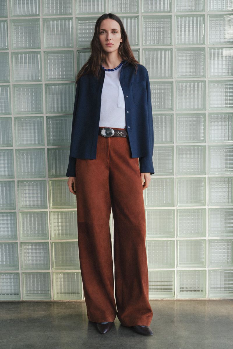 A young adult woman poses in a casual, fashionable outfit consisting of a white t-shirt, an open navy shirt, and rust-colored wide-leg trousers.