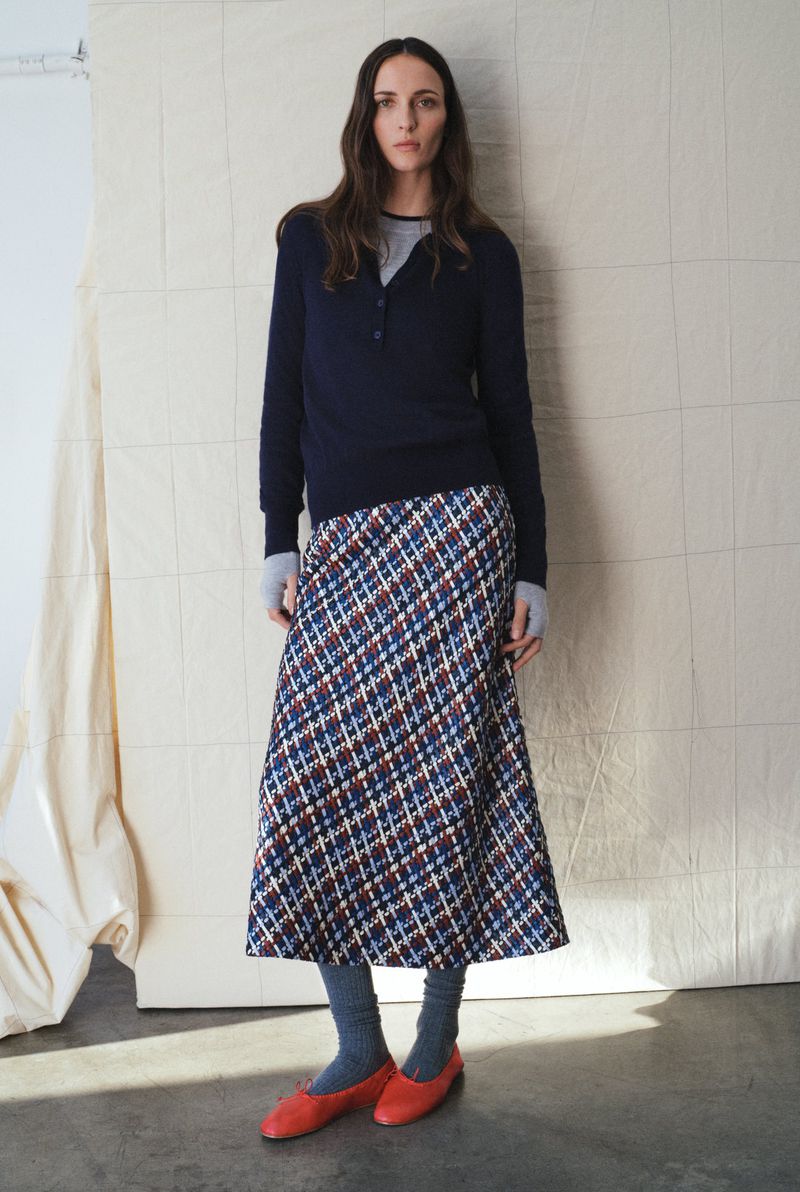 Woman in a studio wearing a navy sweater and plaid skirt with red flats, standing against a light-colored backdrop. Fashion editorial.