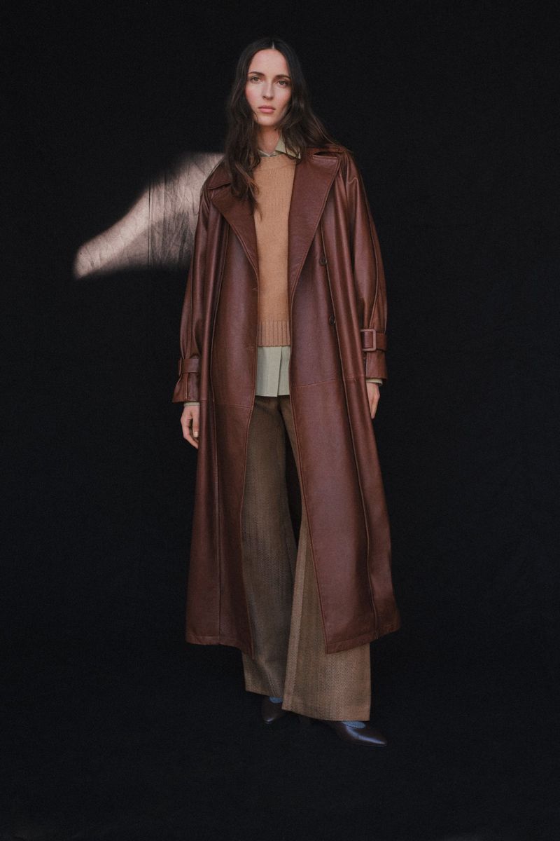 Elegant woman in a brown faux leather trench coat and wide-leg pants strikes a pose against a black background with natural light.