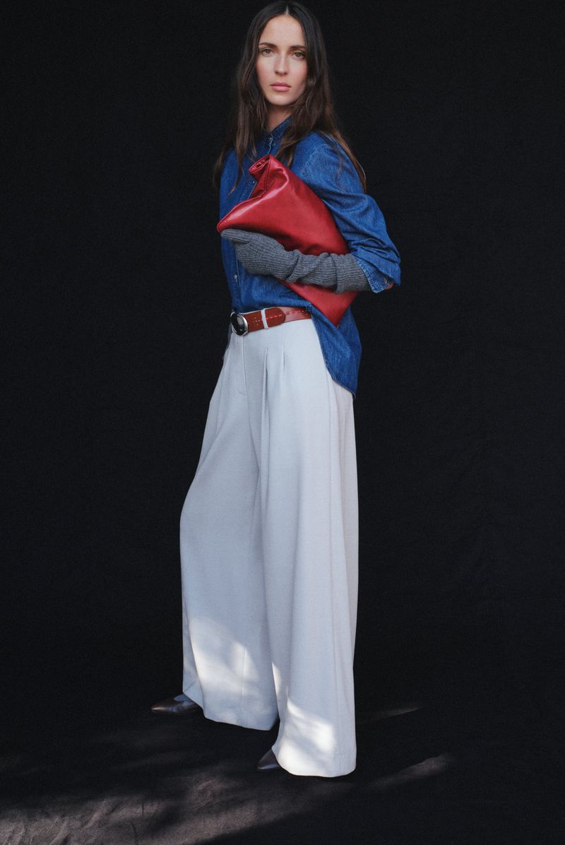 Fashion shot of a woman in a blue denim shirt, white trousers, holding a red clutch, and wearing grey gloves against a dark background.