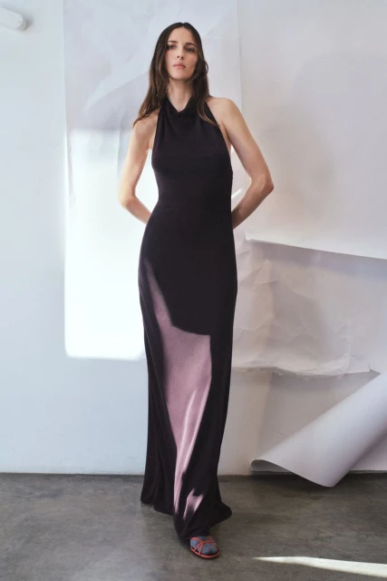 Adult female model posing in a full-length black halter dress with a thigh slit in a studio with neutral background and natural lighting.