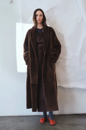 Model Waleska Gorczevski wearing an oversized full-length brown plush coat and red flat shoes against a white backdrop.