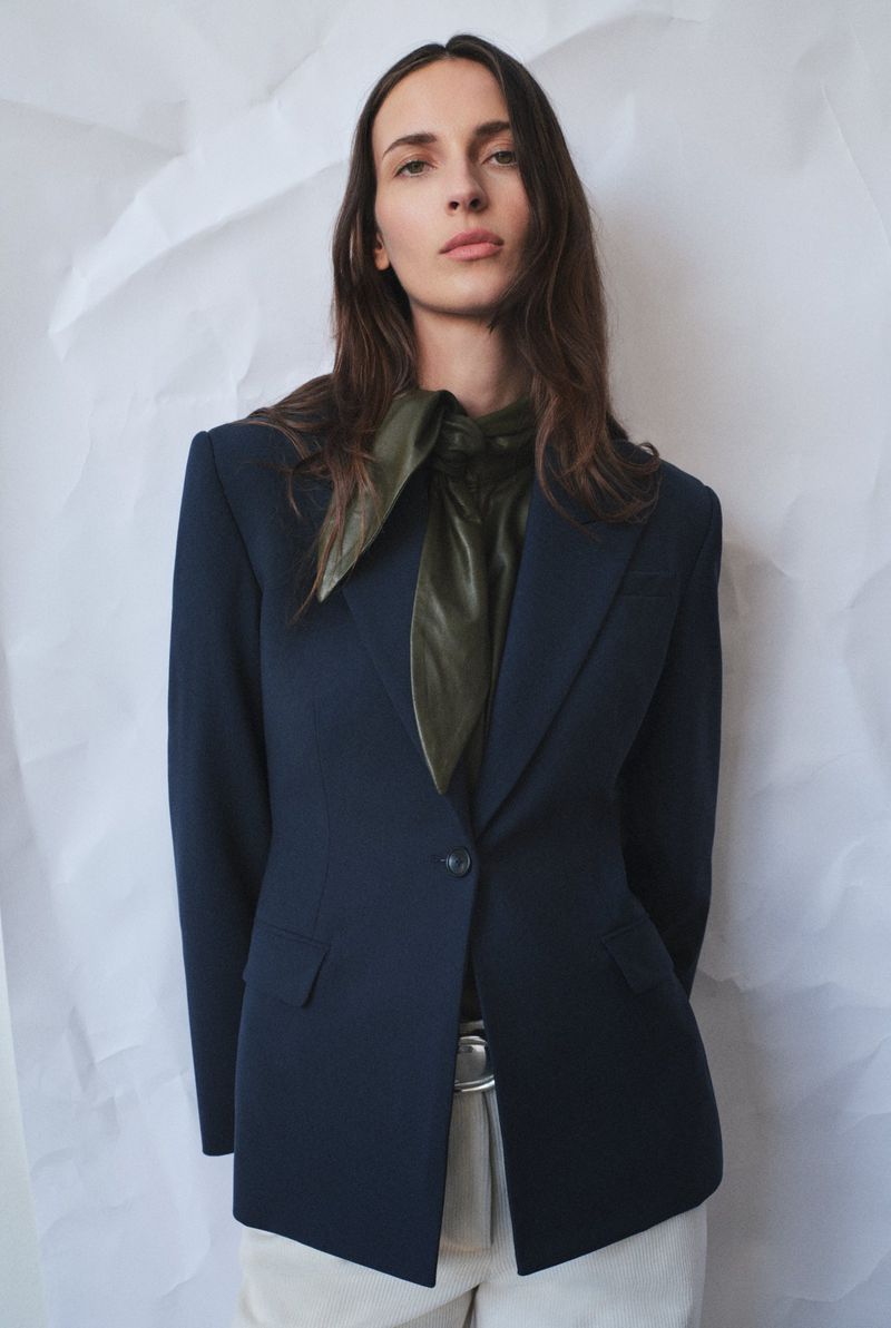 A woman with long brown hair poses in a navy blazer, silky green shirt and white pants against a wrinkled white backdrop.