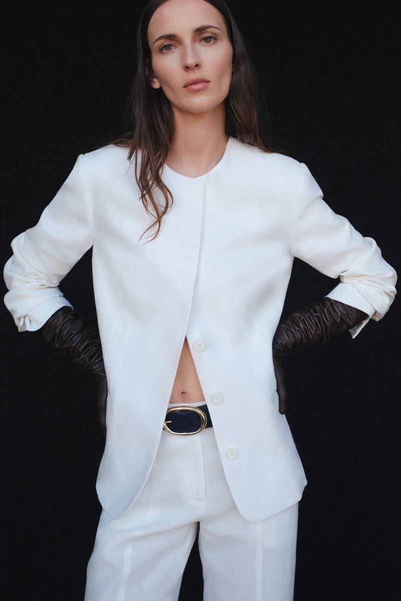 A woman wearing a white linen suit and black gloves poses against a black backdrop, showcasing a stylish and minimalist fashion aesthetic.