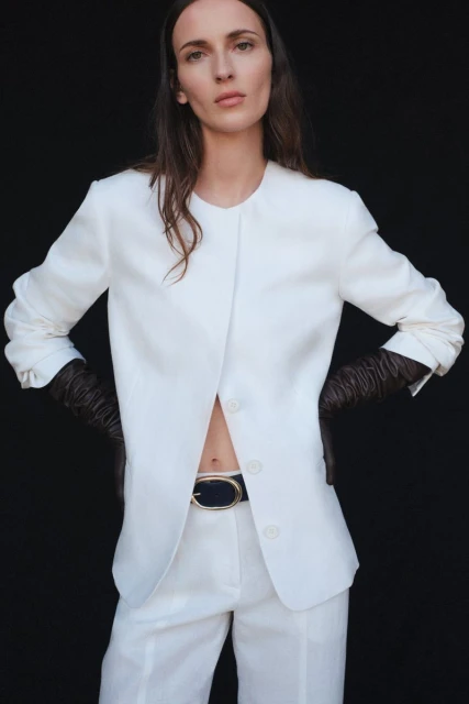 A woman wearing a white linen suit and black gloves poses against a black backdrop, showcasing a stylish and minimalist fashion aesthetic.