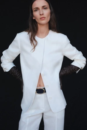 A woman wearing a white linen suit and black gloves poses against a black backdrop, showcasing a stylish and minimalist fashion aesthetic.
