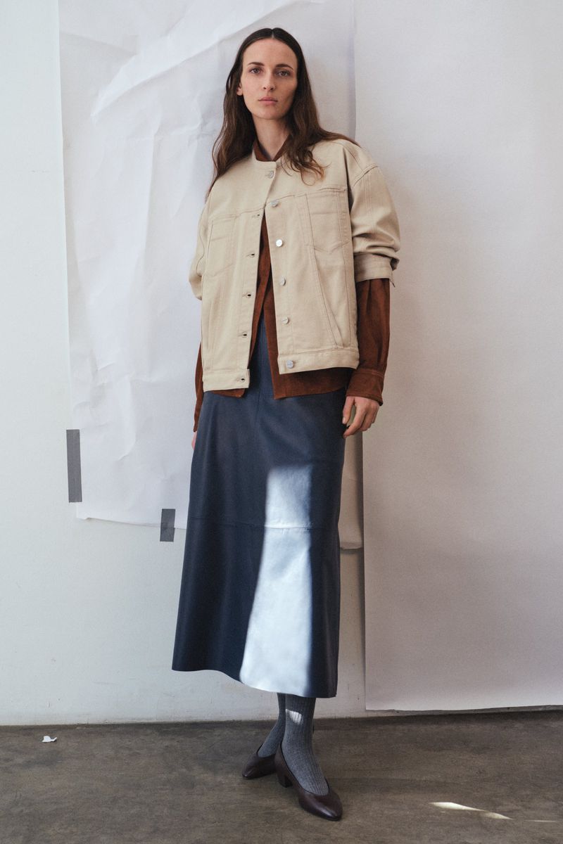 Fashion shot of an adult woman posing indoors, wearing a jacket, long skirt, shirt, stockings, and shoes against a white backdrop.