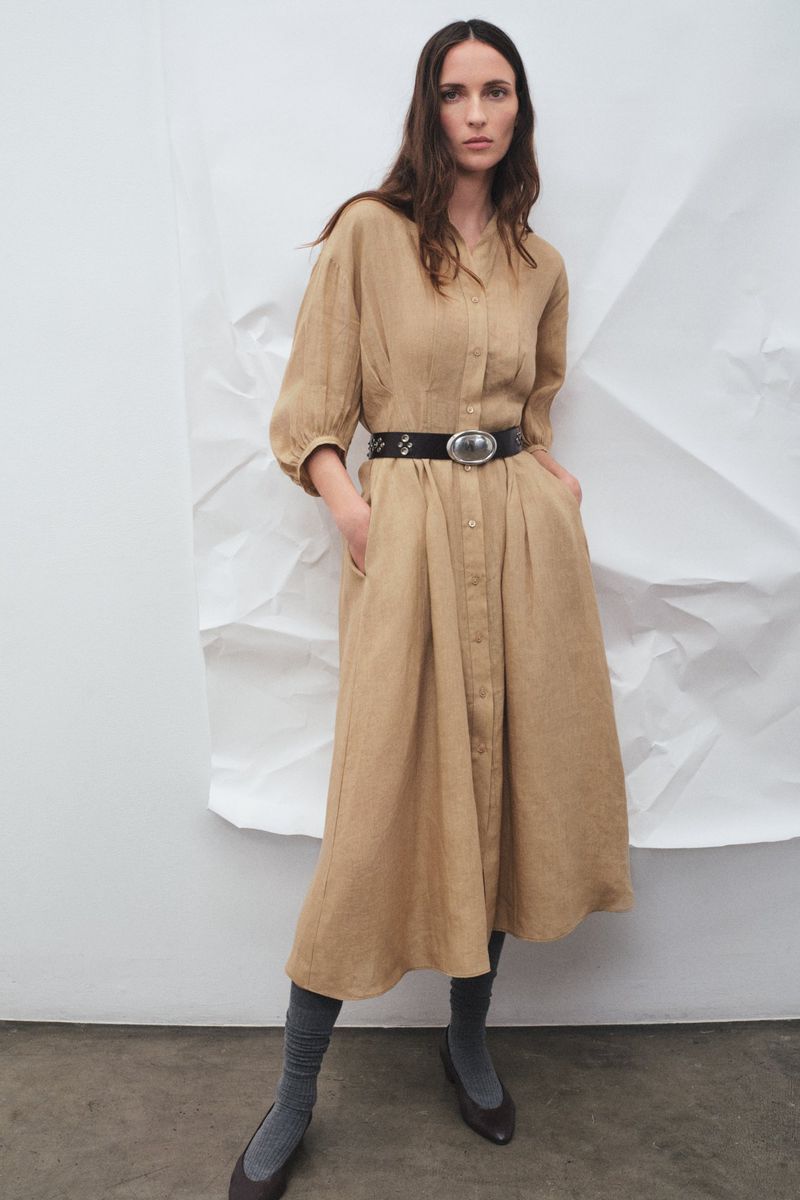 A woman poses in a long-sleeved tan dress with a black belt and gray socks in a studio with a crumpled white backdrop.