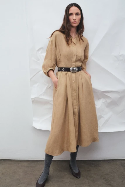 A woman poses in a long-sleeved tan dress with a black belt and gray socks in a studio with a crumpled white backdrop.