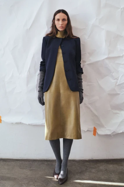 A woman in a dress and jacket stands against a crinkled paper backdrop in a studio setting, showcasing fashion apparel.