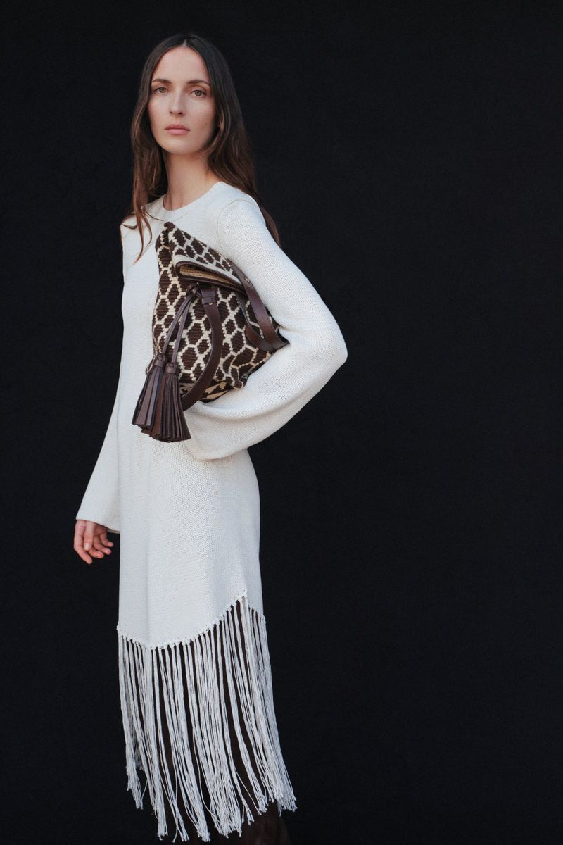An adult woman poses against a black backdrop, wearing a cream-colored fringe dress, and carrying a brown patterned handbag.
