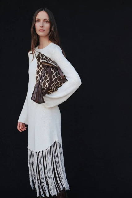 An adult woman poses against a black backdrop, wearing a cream-colored fringe dress, and carrying a brown patterned handbag.