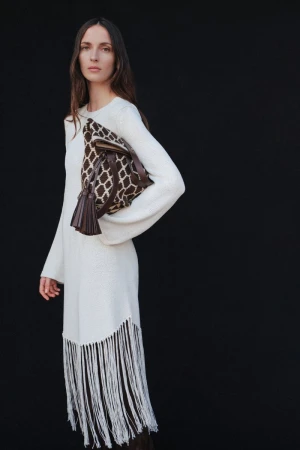 An adult woman poses against a black backdrop, wearing a cream-colored fringe dress, and carrying a brown patterned handbag.