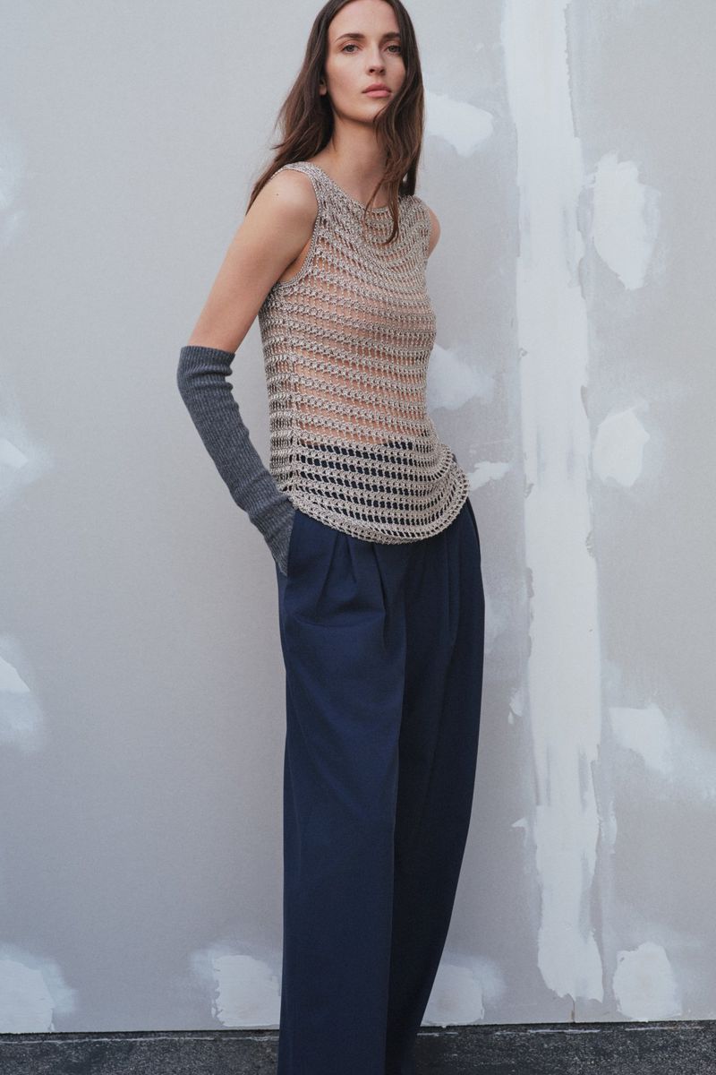 A woman poses in a sleeveless crochet top and navy blue wide-leg pants against a neutral wall, showcasing a minimalist fashion aesthetic.