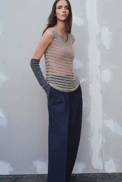 A woman poses in a sleeveless crochet top and navy blue wide-leg pants against a neutral wall, showcasing a minimalist fashion aesthetic.