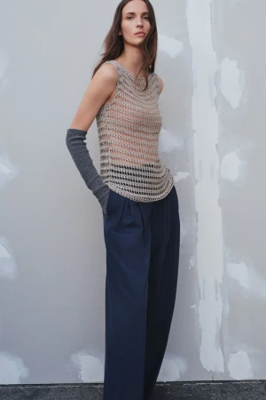 A woman poses in a sleeveless crochet top and navy blue wide-leg pants against a neutral wall, showcasing a minimalist fashion aesthetic.