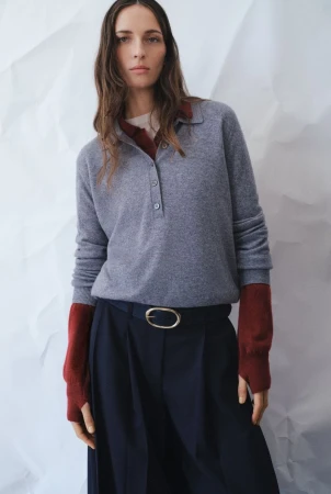 Fashion portrait of an adult female wearing a grey polo shirt and navy pants, posing in front of a white backdrop. Studio lighting.