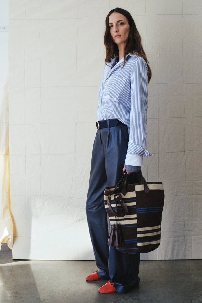 Waleska Gorczevski wears a blue striped button-down shirt, navy wide-leg trousers, and red shoes while holding a striped tote bag.