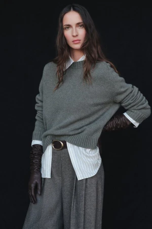 Waleska Gorczevski poses in an olive green knit sweater, striped shirt, pleated trousers, and leather gloves against a black backdrop.