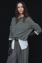 Waleska Gorczevski poses in an olive green knit sweater, striped shirt, pleated trousers, and leather gloves against a black backdrop.