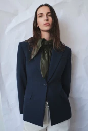Waleska Gorczevski wearing a tailored navy blazer and green neck scarf against a crinkled white paper backdrop.