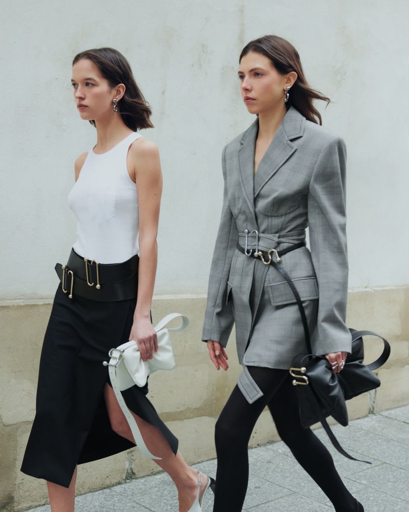 Two women in stylish outfits walk against a white wall, one in a white top and black skirt, the other in a grey blazer and black tights.
