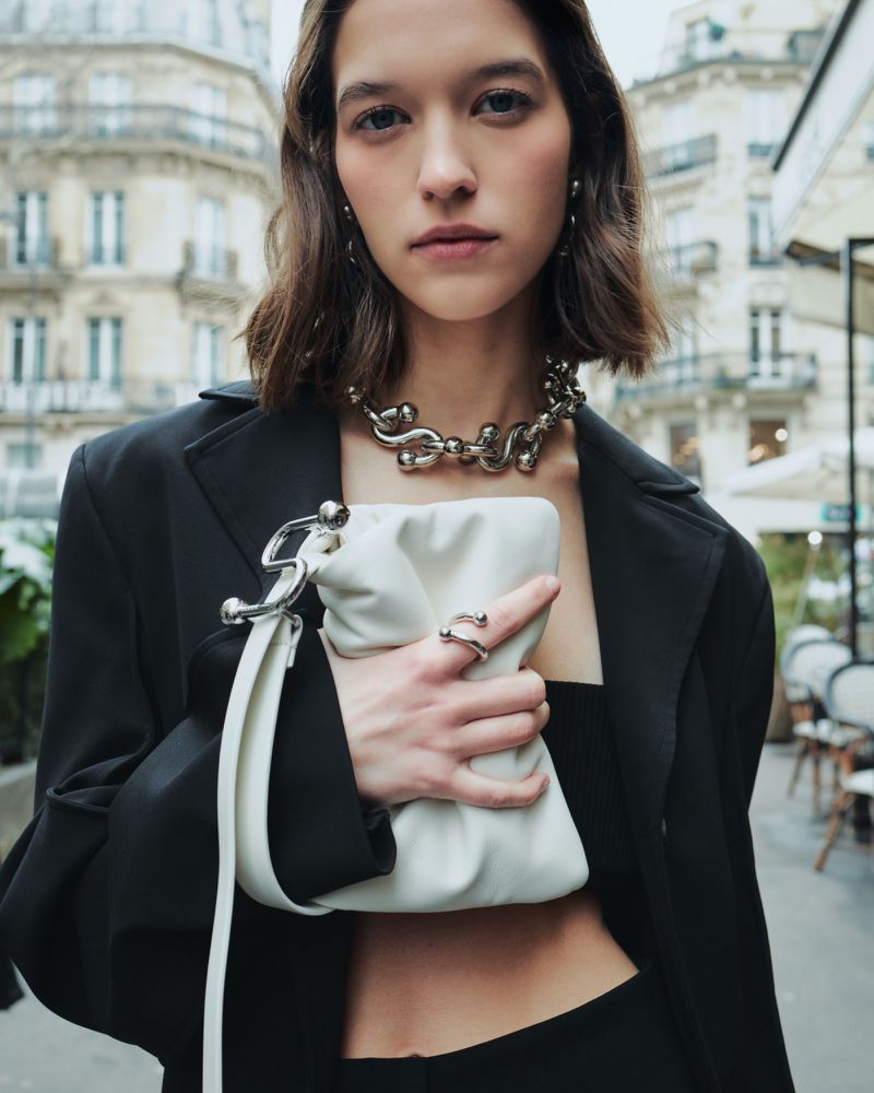 Model Léa Julian holds a white leather clutch with silver hardware, wearing a black blazer and bold silver chain necklace on a city street.