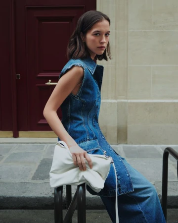 Model Léa Julian in a sleeveless denim dress holding a white leather bag while leaning against a street railing by a red doorway.