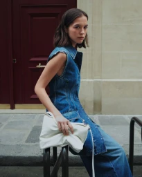 Model Léa Julian in a sleeveless denim dress holding a white leather bag while leaning against a street railing by a red doorway.