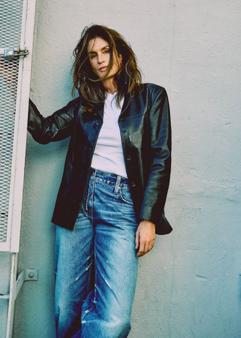 Stylish woman with long brown hair poses in jeans and black jacket against grey wall with metal fixture in an urban setting.