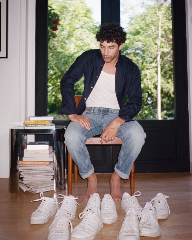 A man sitting on a chair indoors surrounded by several pairs of white sneakers, wearing casual clothing with a window behind him.